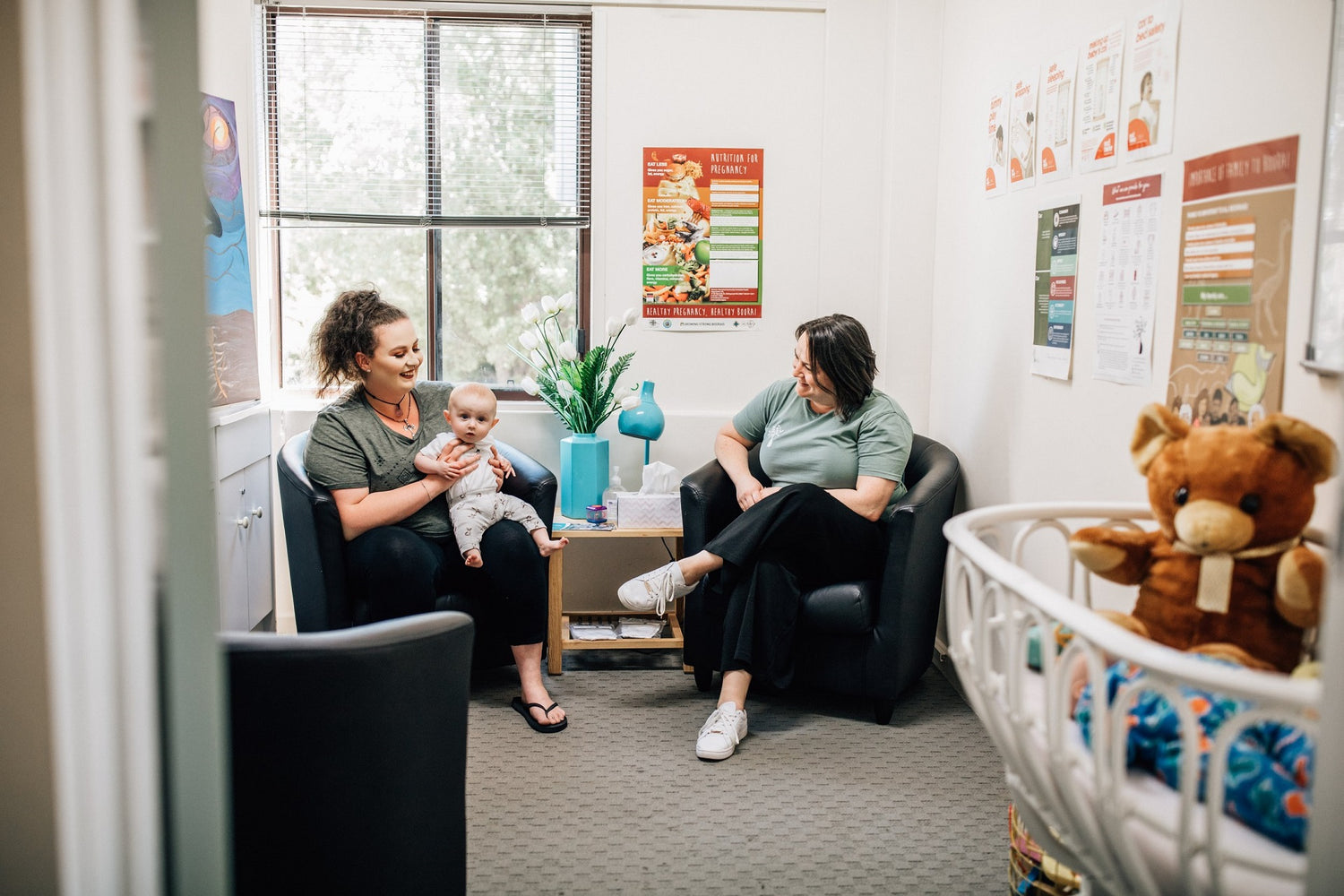 A family support worker is sitting in a bright room with a mum and her baby. They are both smiling and the baby is looking at the camera. There is a teddy bear in a bassinet on the right hand side of the image.