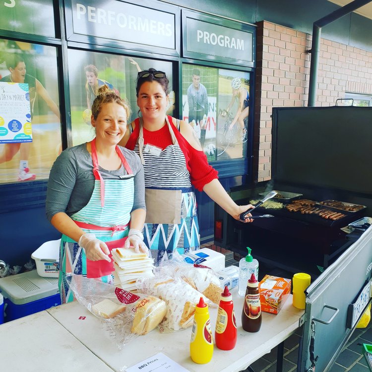 Two smiling volunteers are looking at the camera. They are at a BBQ with sausages, onion and bread and are doing a sausage sizzle.