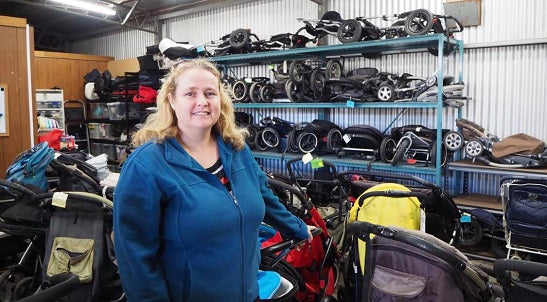 A volunteer of Olivia's Place is standing with lots of prams behind her. The volunteer is looking at the camera and smiling and she has her hand on one of the prams